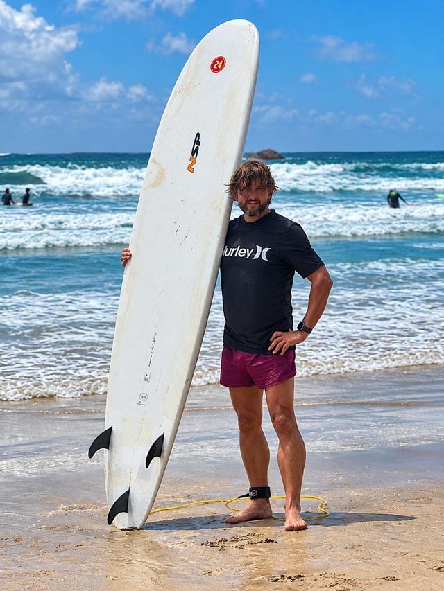 Gary Collins smiling on a beach, standing beside a white surfboard